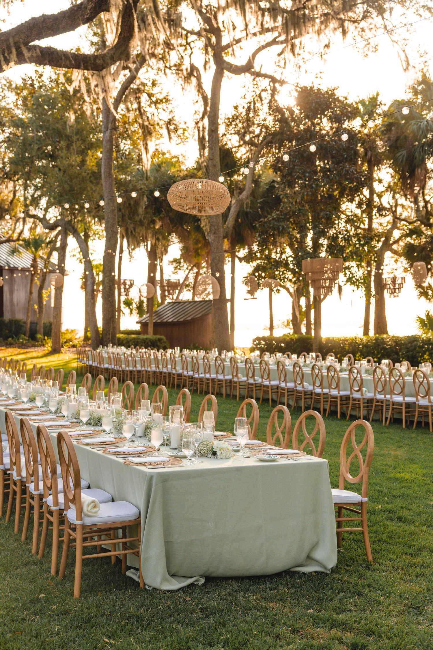 Elegant outdoor wedding reception setup with white-clothed tables and wooden chairs under oak trees.