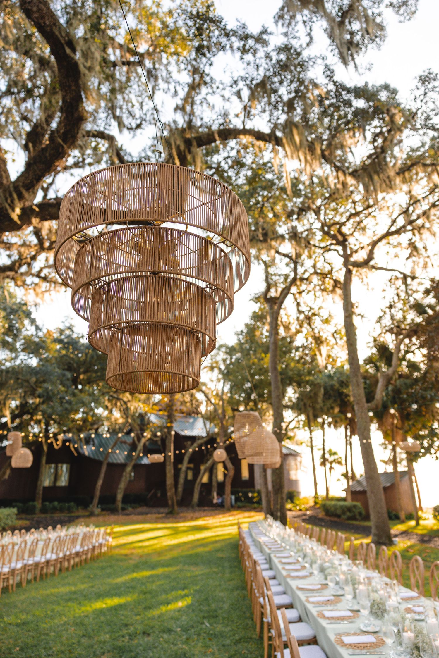 Elegant outdoor wedding reception setup with woven basket pendant lights hanging over long dining tables under oak trees.
