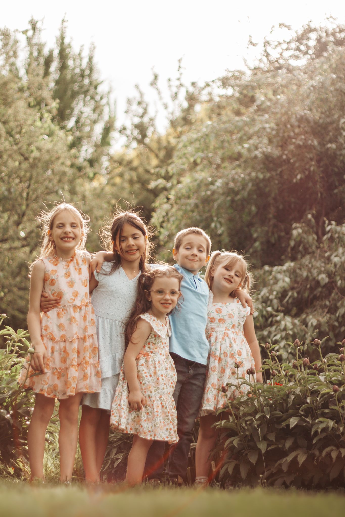 Children in summer outfits pose together against a garden backdrop during golden hour lighting.