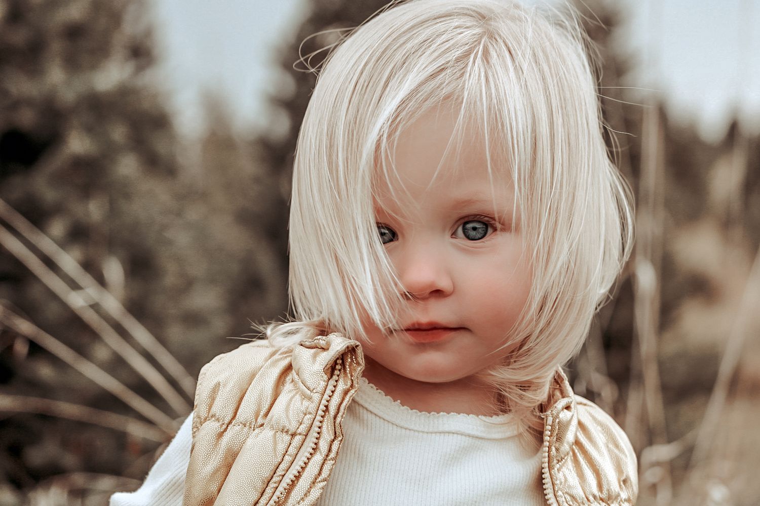 A young child with platinum blonde hair wearing a beige sweater and white shirt in soft natural lighting.