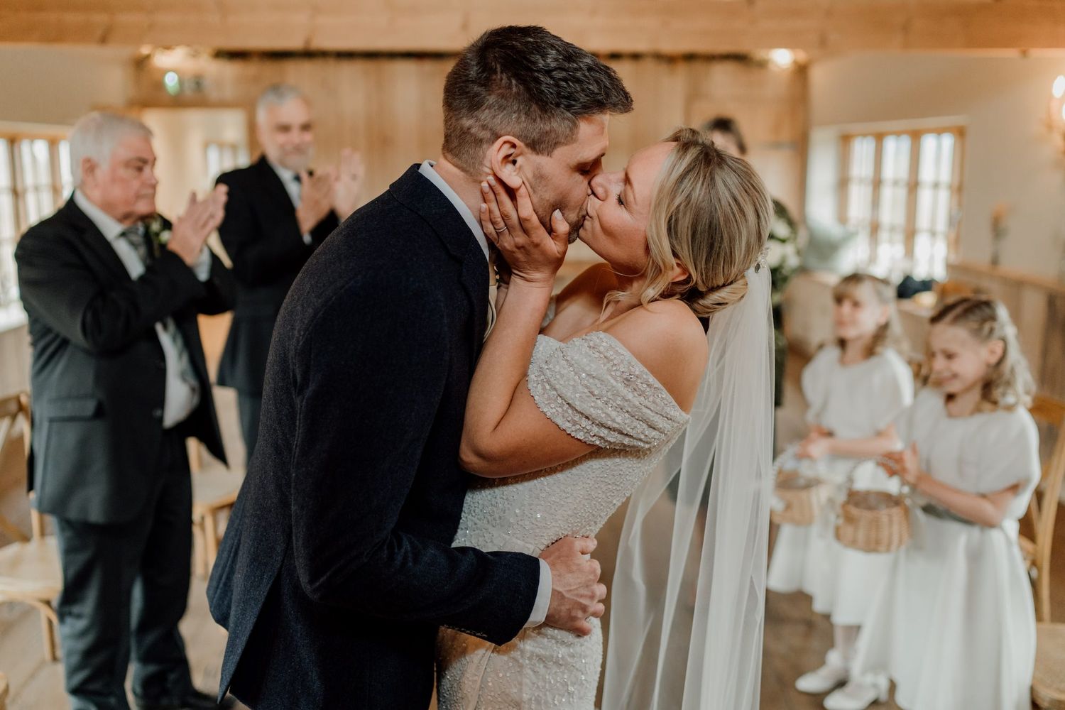 Wedding couple shares romantic kisses in a light-filled venue with elegant decor and guests in formal attire.