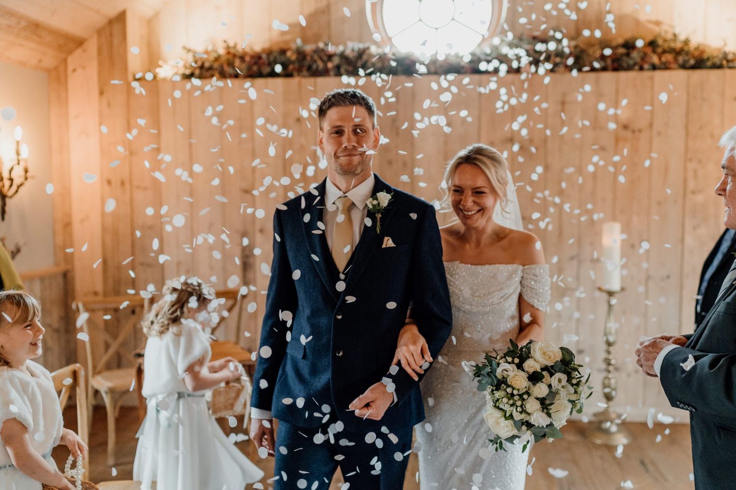 White confetti falls over a festive indoor wedding celebration with decorative lighting and floral arrangements.