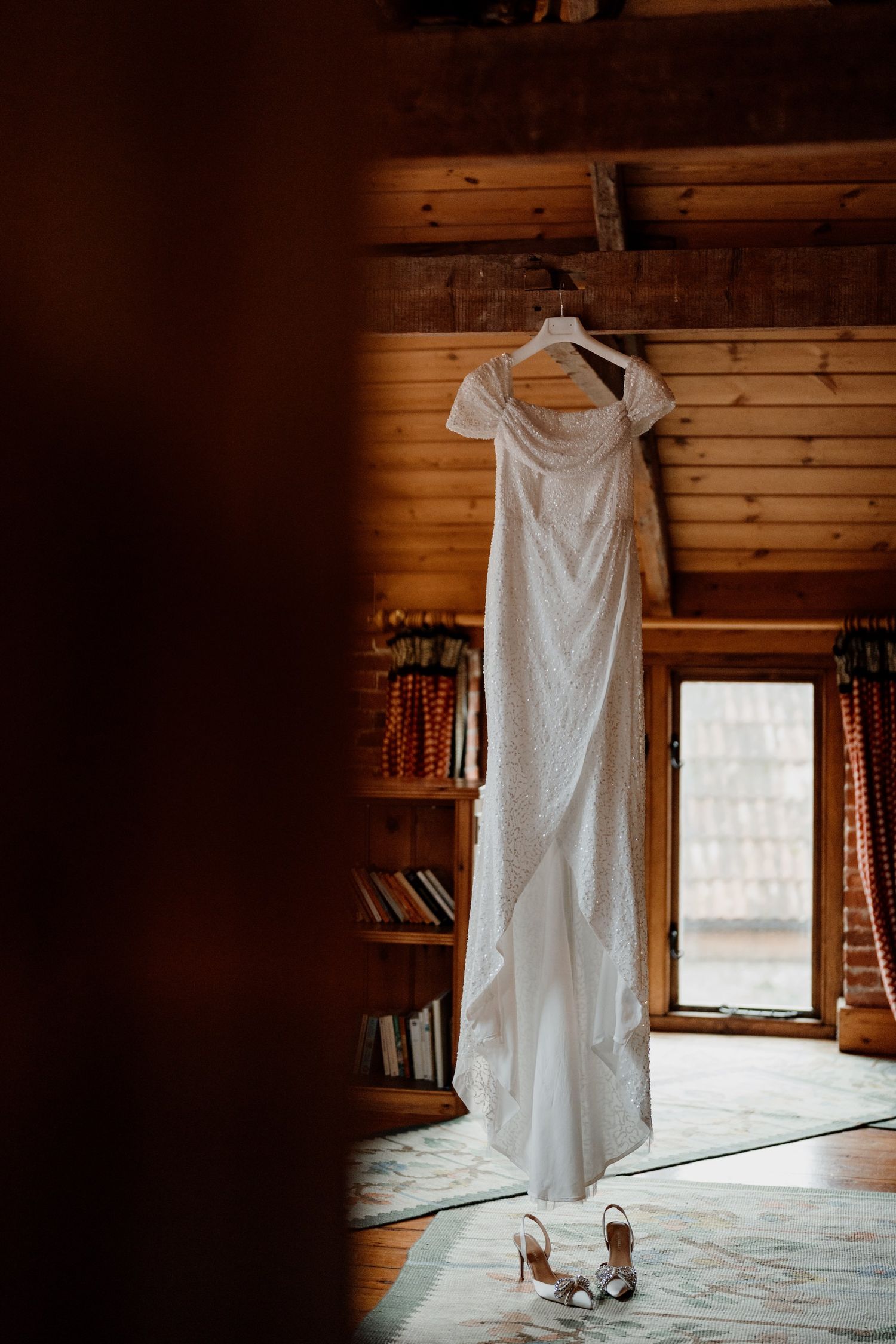 A white wedding dress hangs in a rustic cabin interior with wooden walls and natural window light.