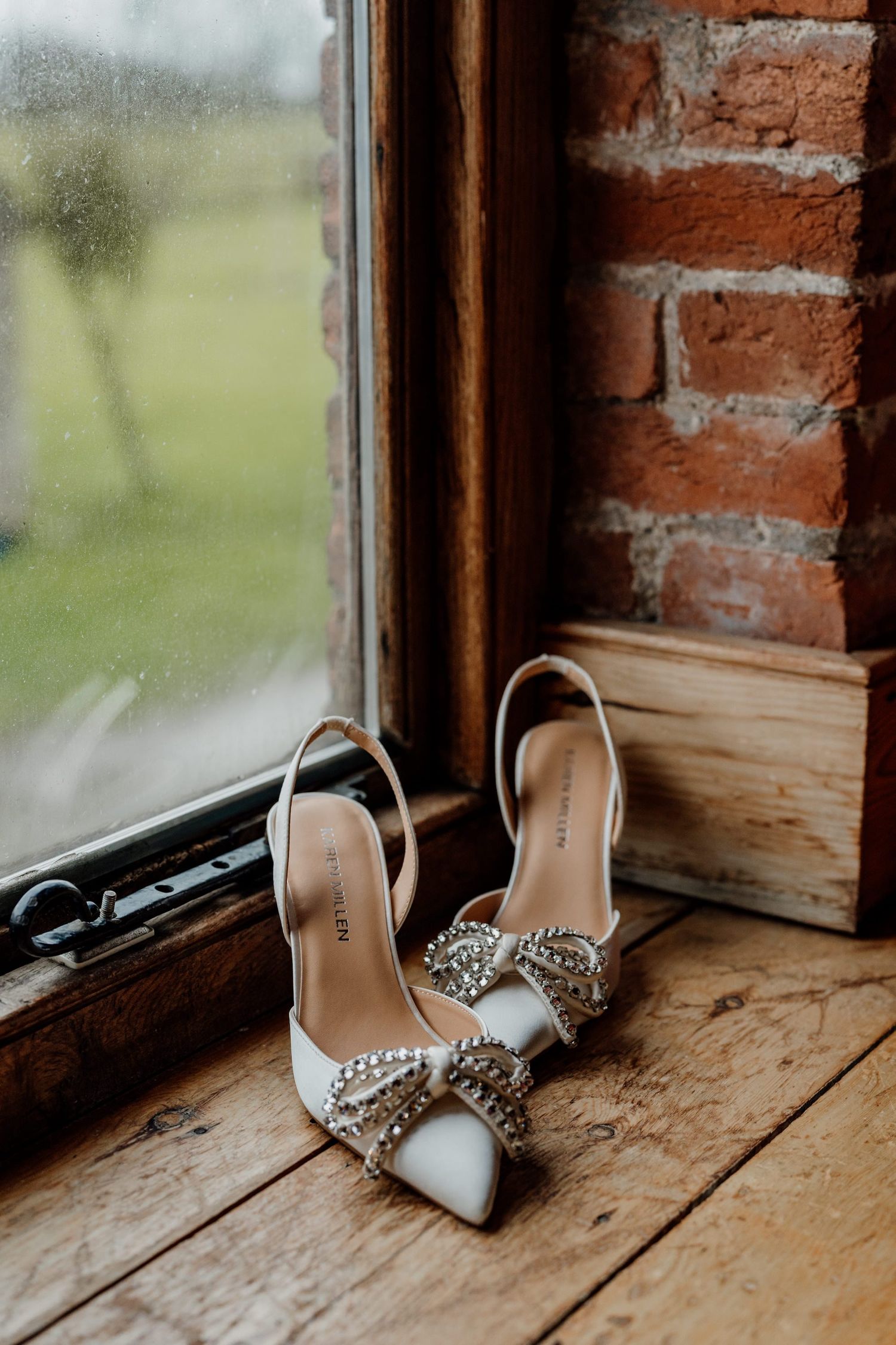 Elegant white bridal shoes with crystal embellishments displayed on a rustic wooden windowsill.