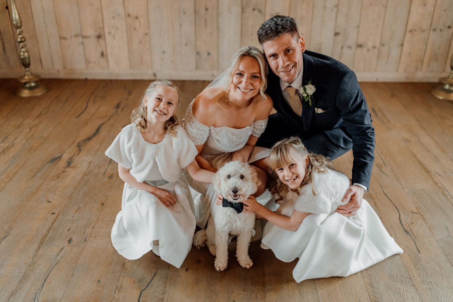 White-dressed group poses playfully on wooden floor with small dog in rustic indoor setting.