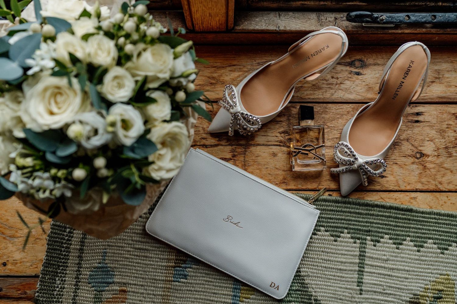 Wedding accessories including silver shoes, white clutch and rose bouquet arranged on wooden floor.