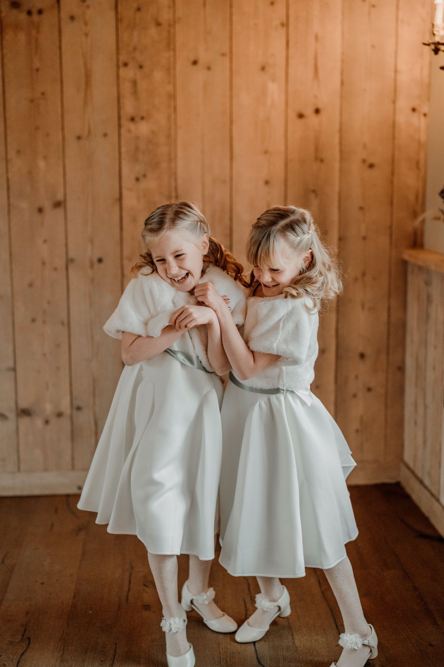 Flower girls in matching white dresses and socks giggle together on wooden floor during wedding celebration.