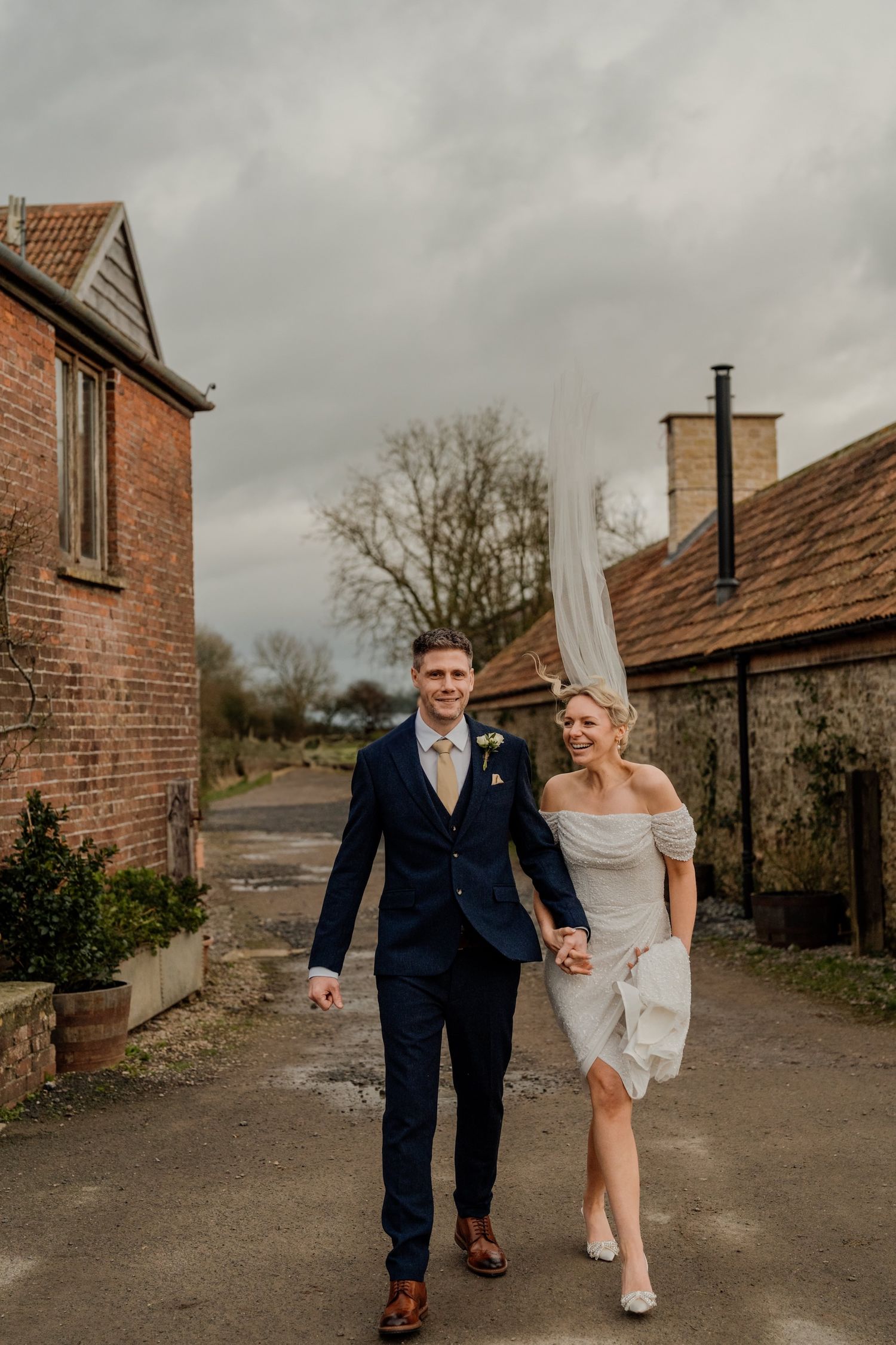 A couple walks hand in hand along a rustic brick path between countryside buildings on their wedding day.
