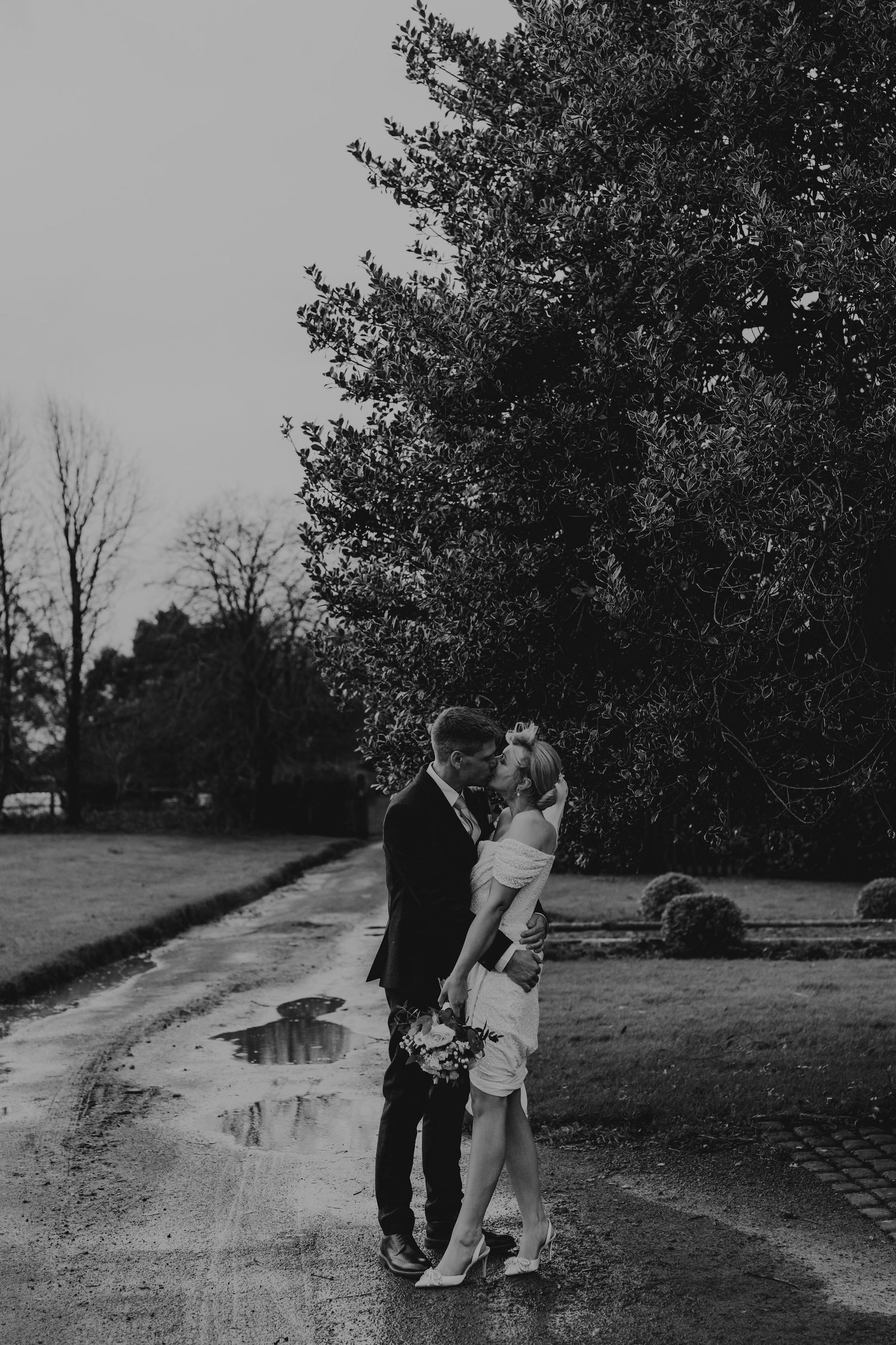 Black and white photo of a couple walking through a countryside path on a cloudy day.