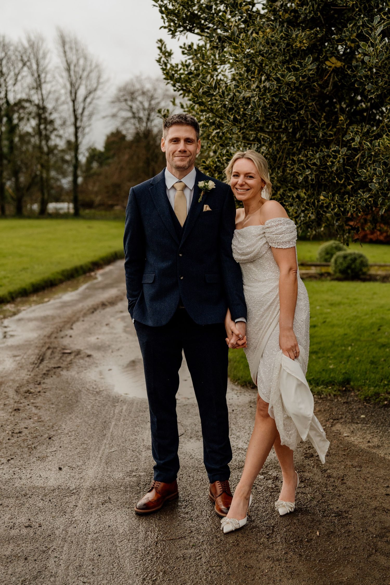 A couple poses together on a gravel path with green gardens and trees in the background.