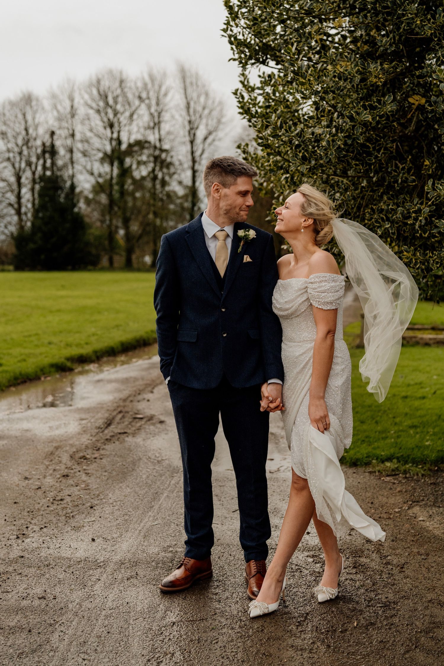 Wedding couple walks together on country path, bride's veil flowing in winter landscape.