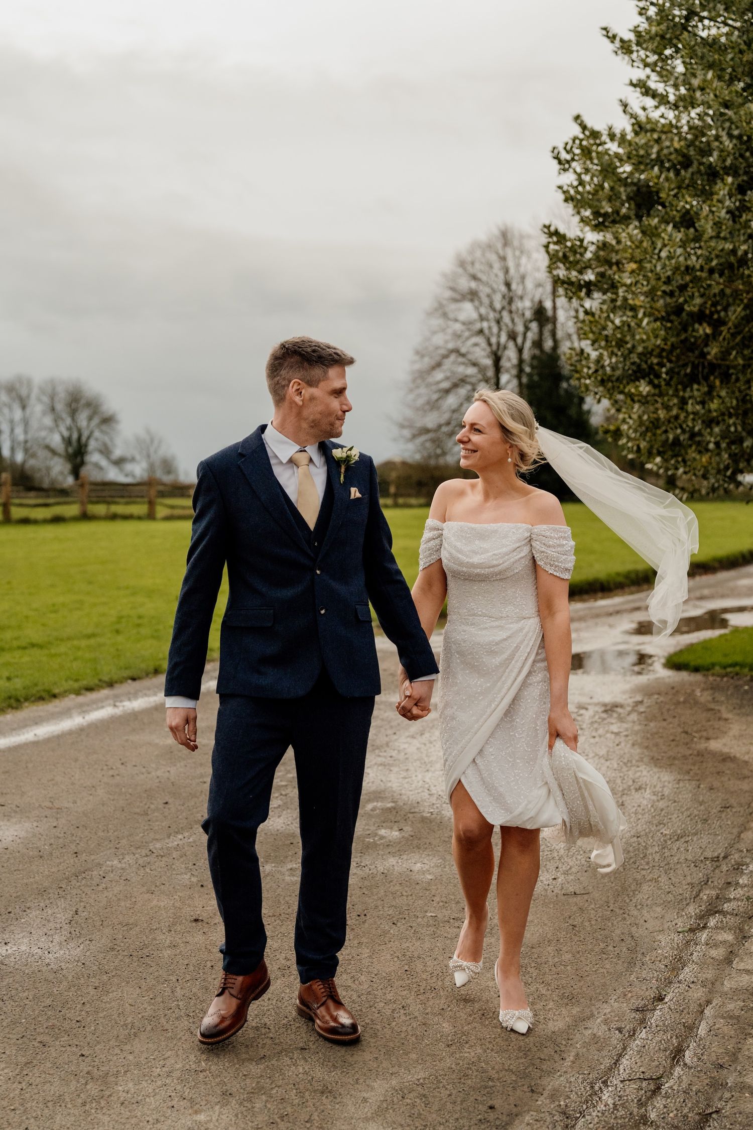 A couple walking hand in hand along a countryside path with a flowing wedding veil in the breeze.