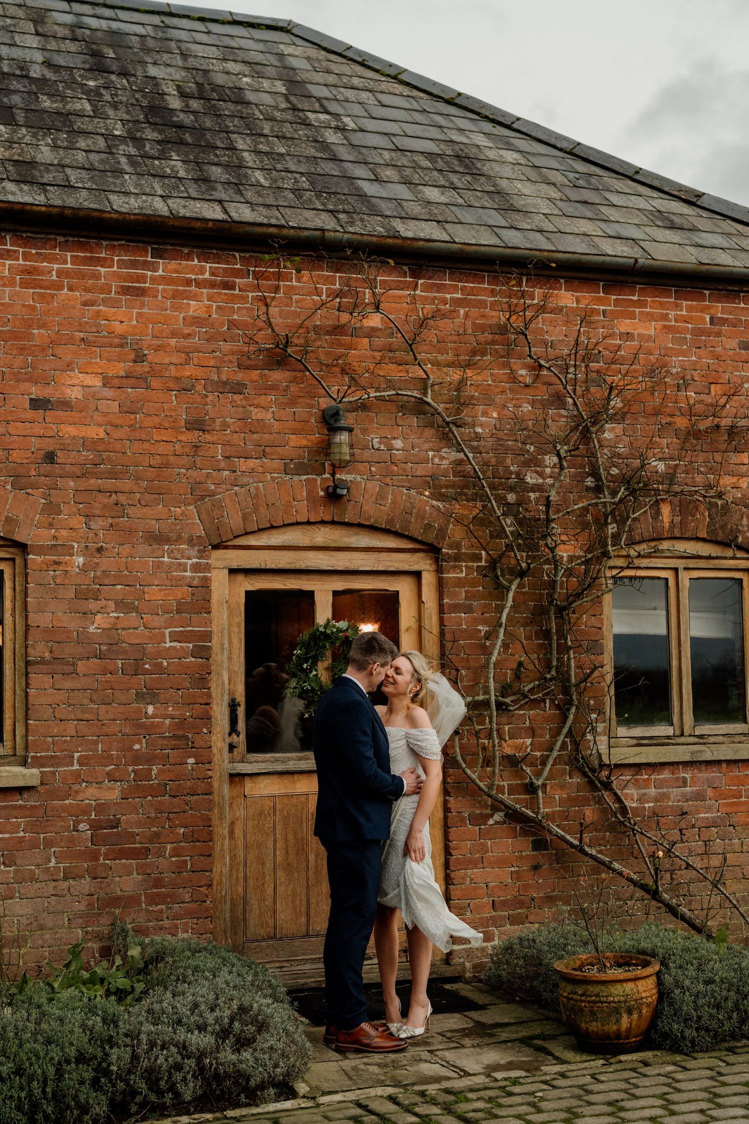 A couple shares a romantic moment against a rustic brick building exterior with climbing vines.