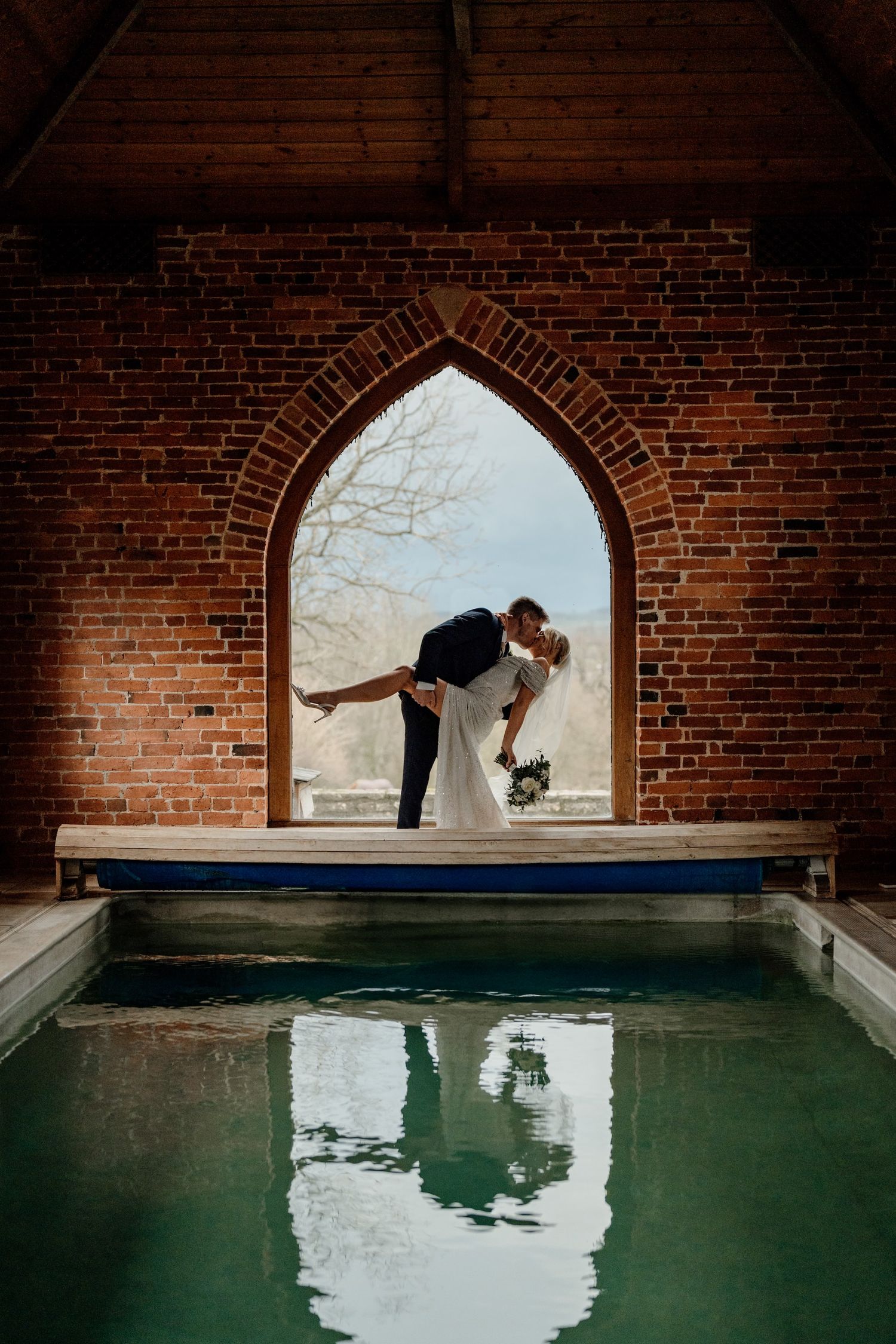 A couple dancing gracefully in an arched brick window above a reflective indoor pool creates a romantic silhouette effect.