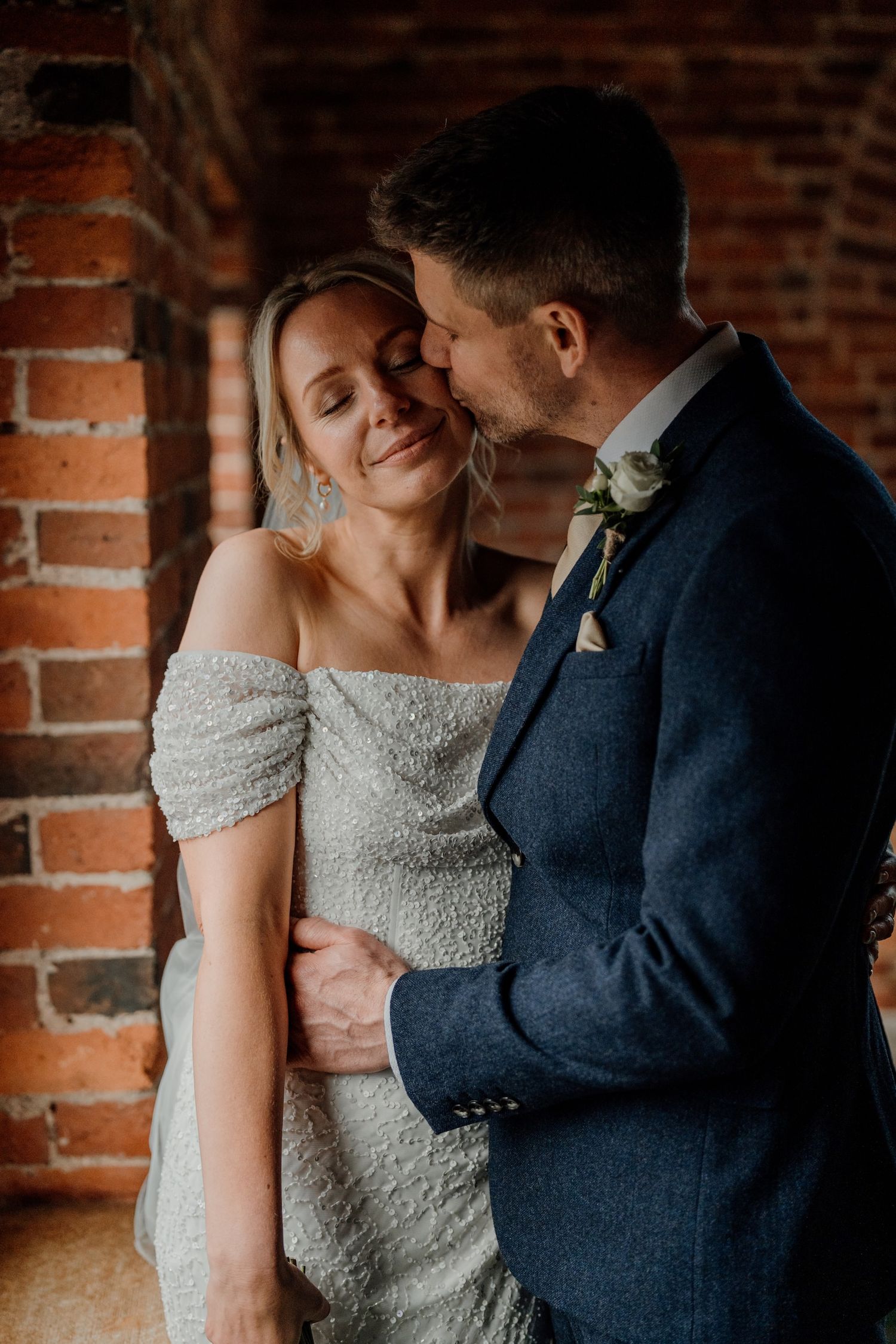 A bride in an off-shoulder dress and groom in navy suit share an intimate moment against exposed brick walls.