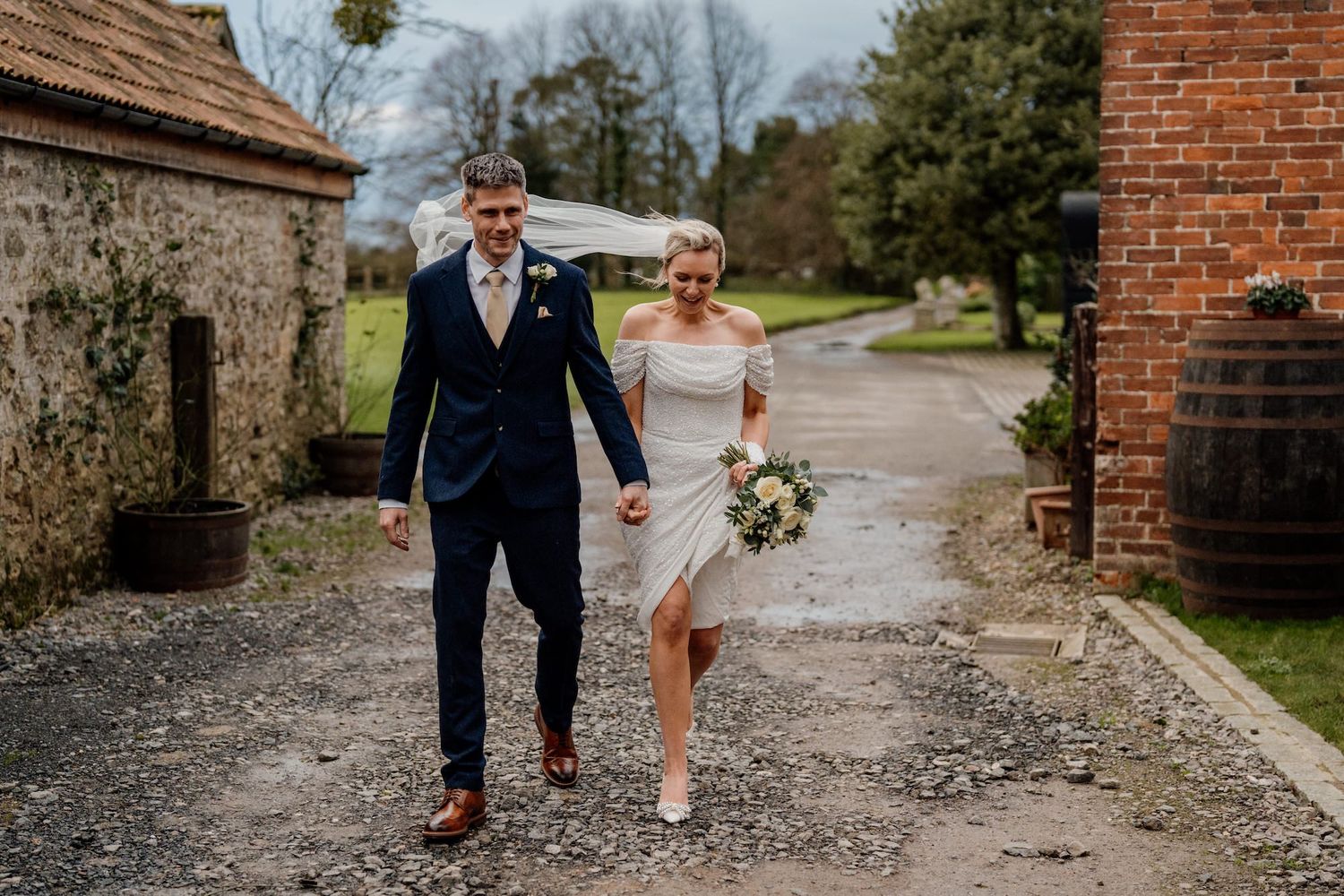 A couple walking together through the grounds of a historic brick building on their wedding day.