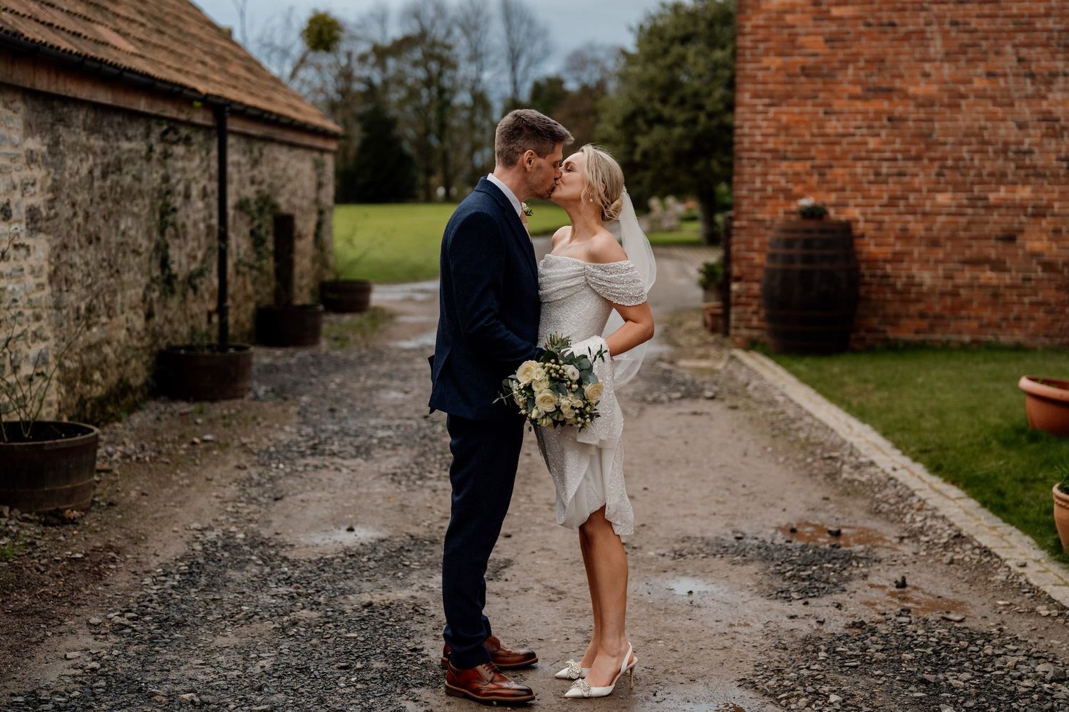 Newlywed couple strolling along a gravel path beside rustic brick buildings.