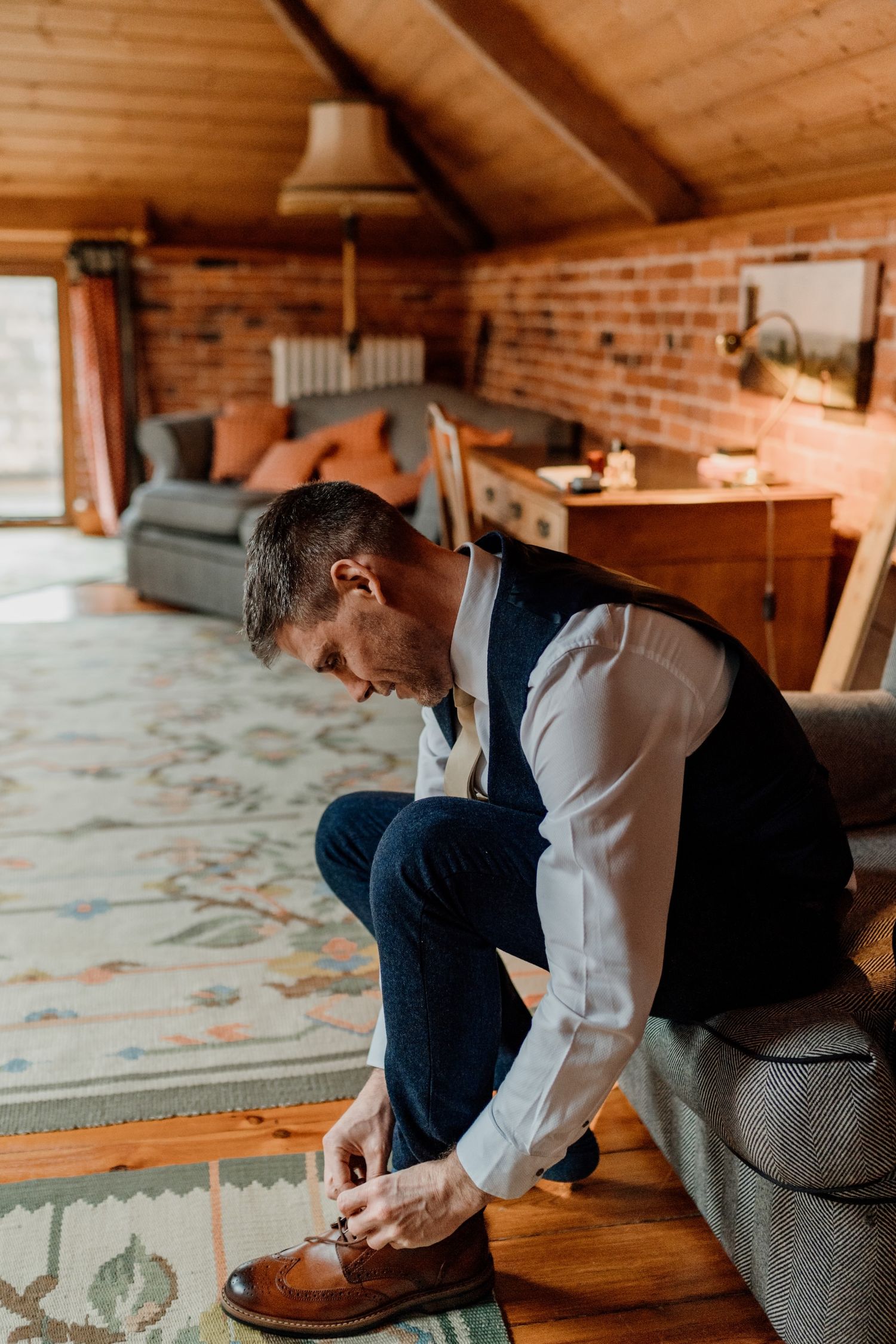 Person getting dressed in formal attire, putting on brown leather dress shoes in a rustic wooden room setting.