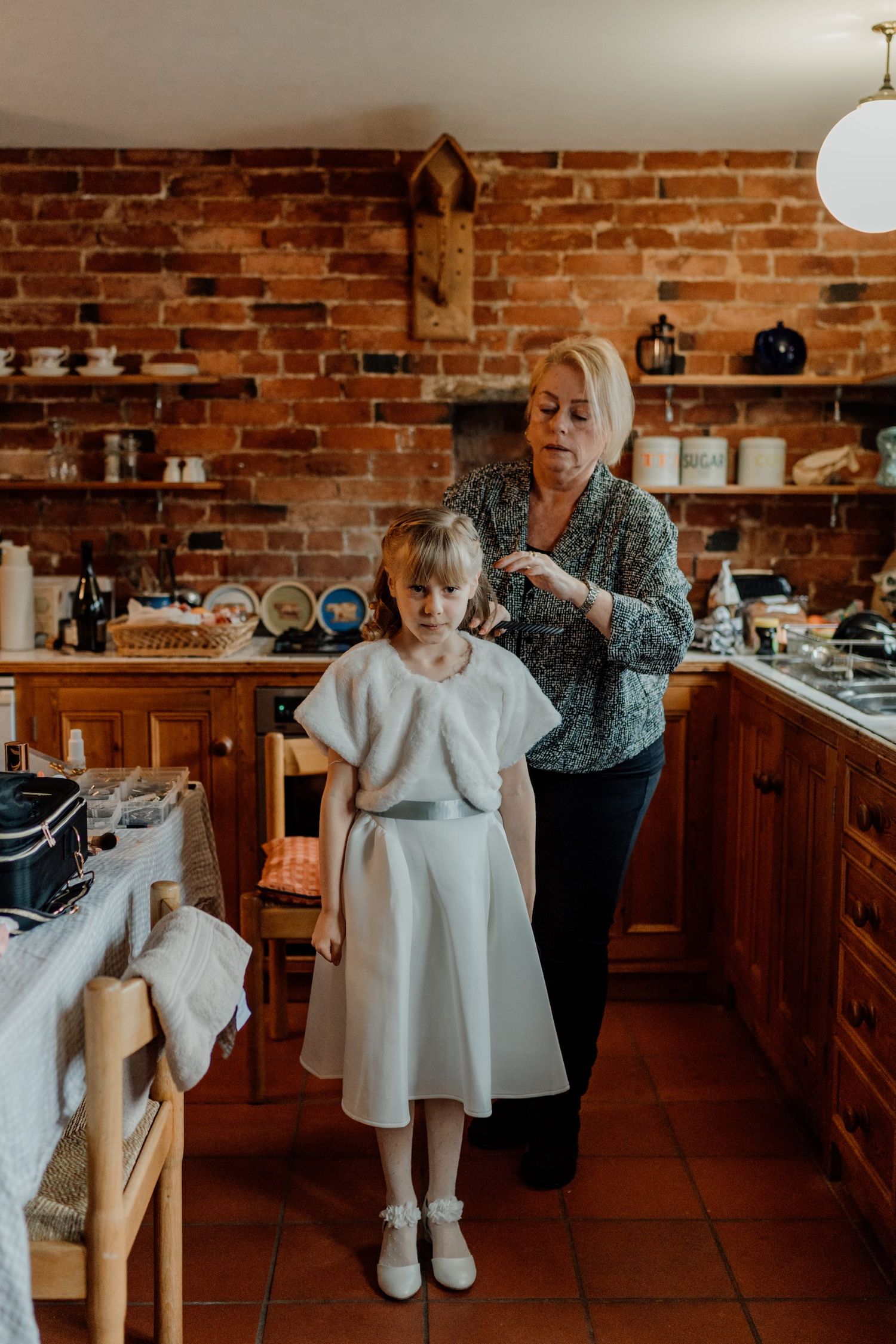 Young flower girls in white dresses sit in a rustic kitchen with exposed brick walls.