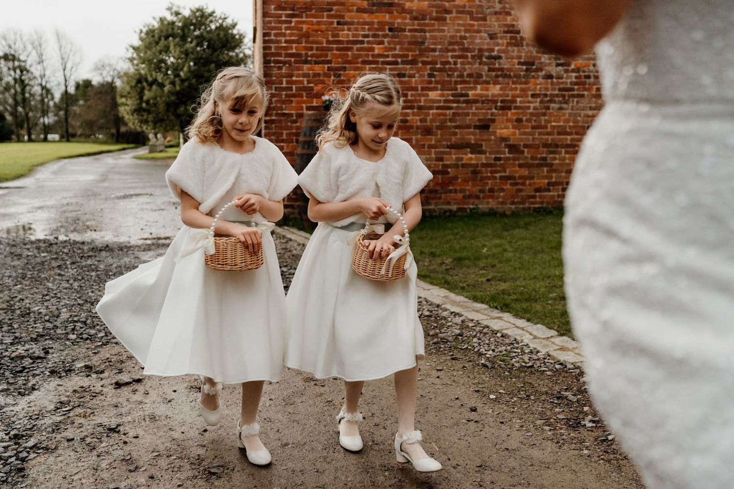 Small children in white dresses walking along a gravel path outdoors.