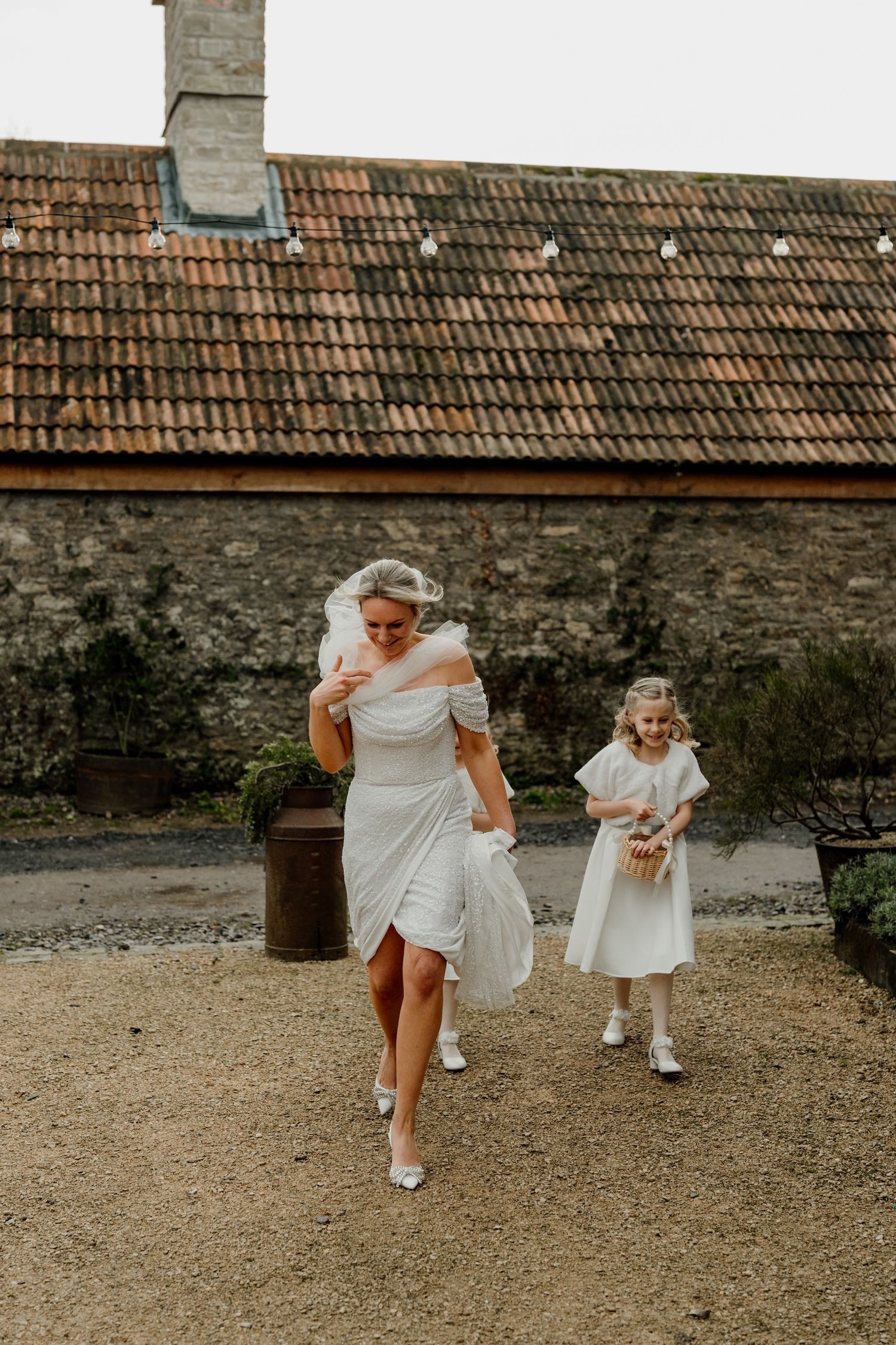 Two people in white outfits run playfully across a gravel courtyard near stone walls and tiled roofs.