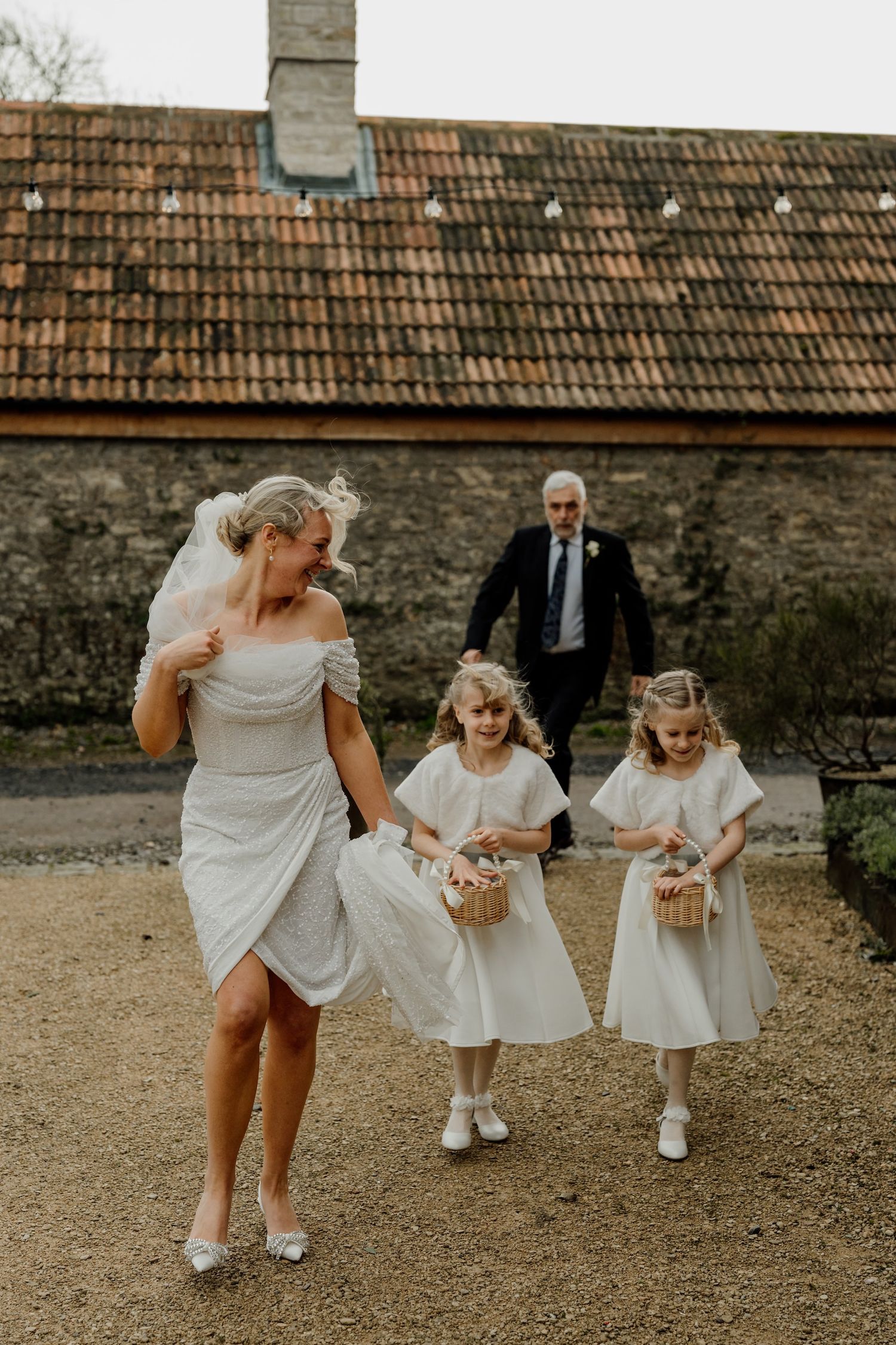Young flower girls in white dresses and matching shoes play on stone gravel path.