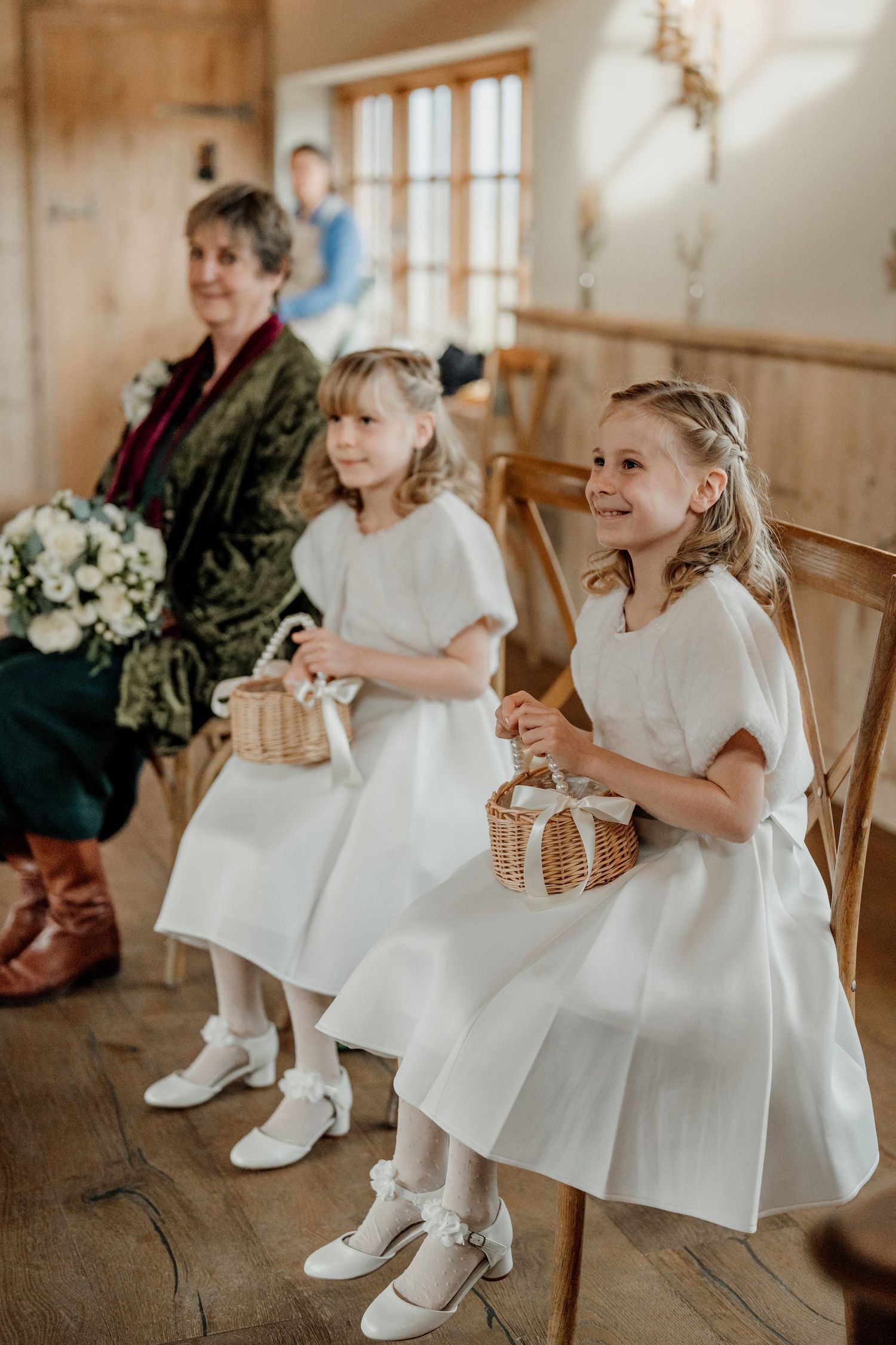 Flower girls in white dresses and boots sit together on wooden chairs.