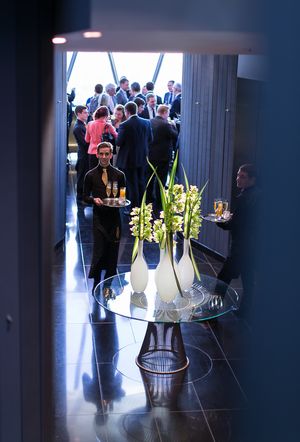 Servers carry drinks through an elegant event space with a glass table displaying floral arrangements in the foreground.