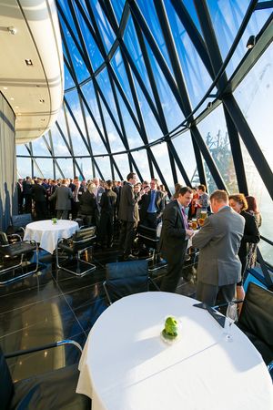 A corporate event or reception in a modern venue with geometric glass windows and guests mingling around white-clothed tables.