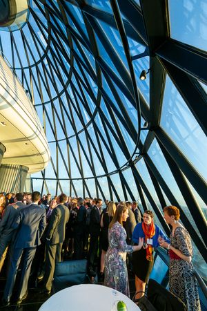 Group of people gather at a modern venue with dramatic angular glass windows and blue sky views during a social event.