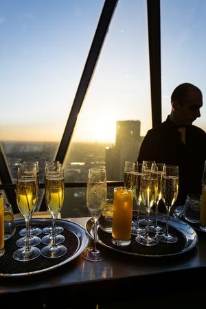 Silhouetted champagne glasses and drinks line a window ledge against a sunset cityscape view.