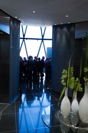 Silhouettes of people stand near large diagonal windows in a modern office building with reflective blue floors and dark walls.