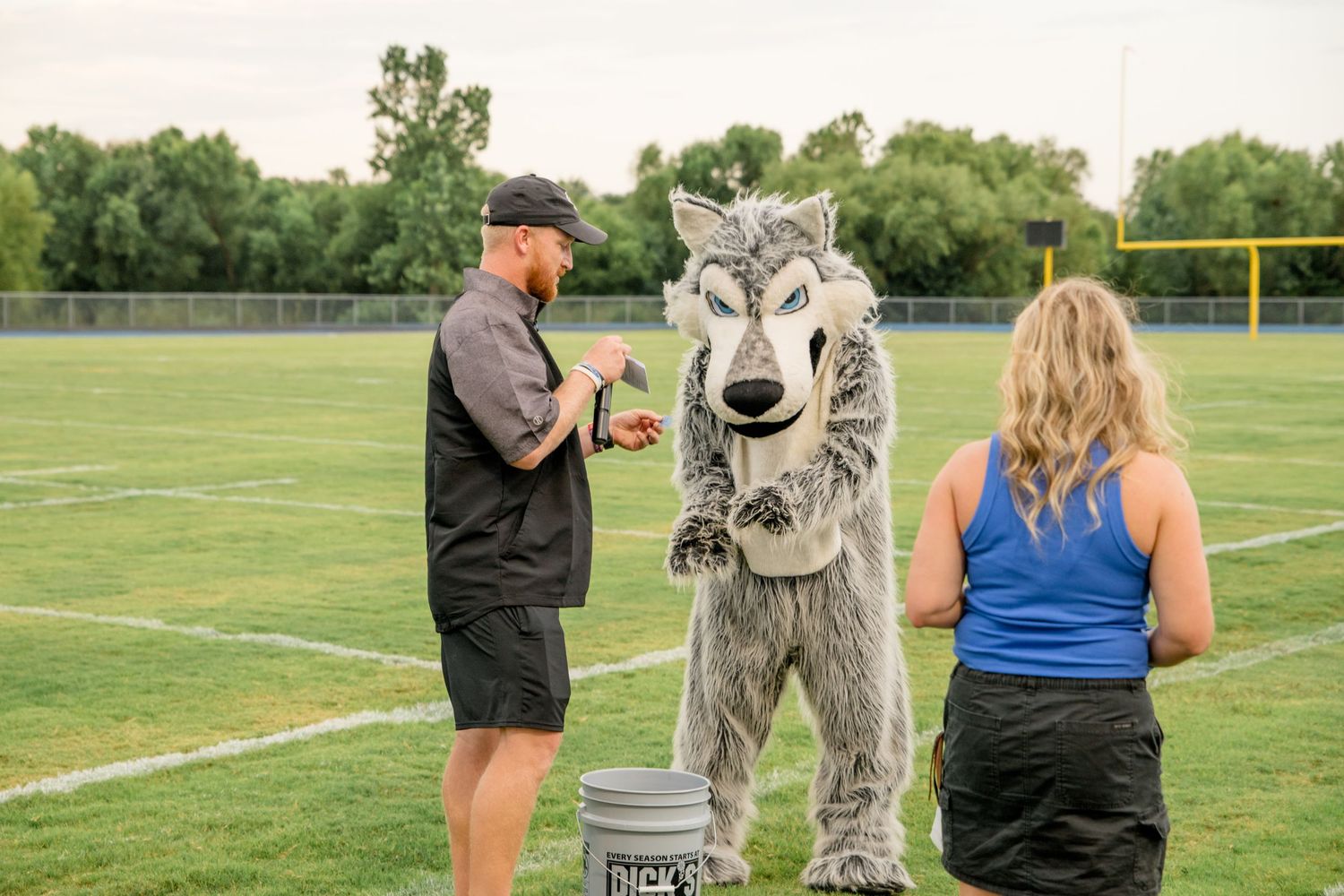 A school mascot character in a wolf costume interacts with people on an athletic field with trees in the background.