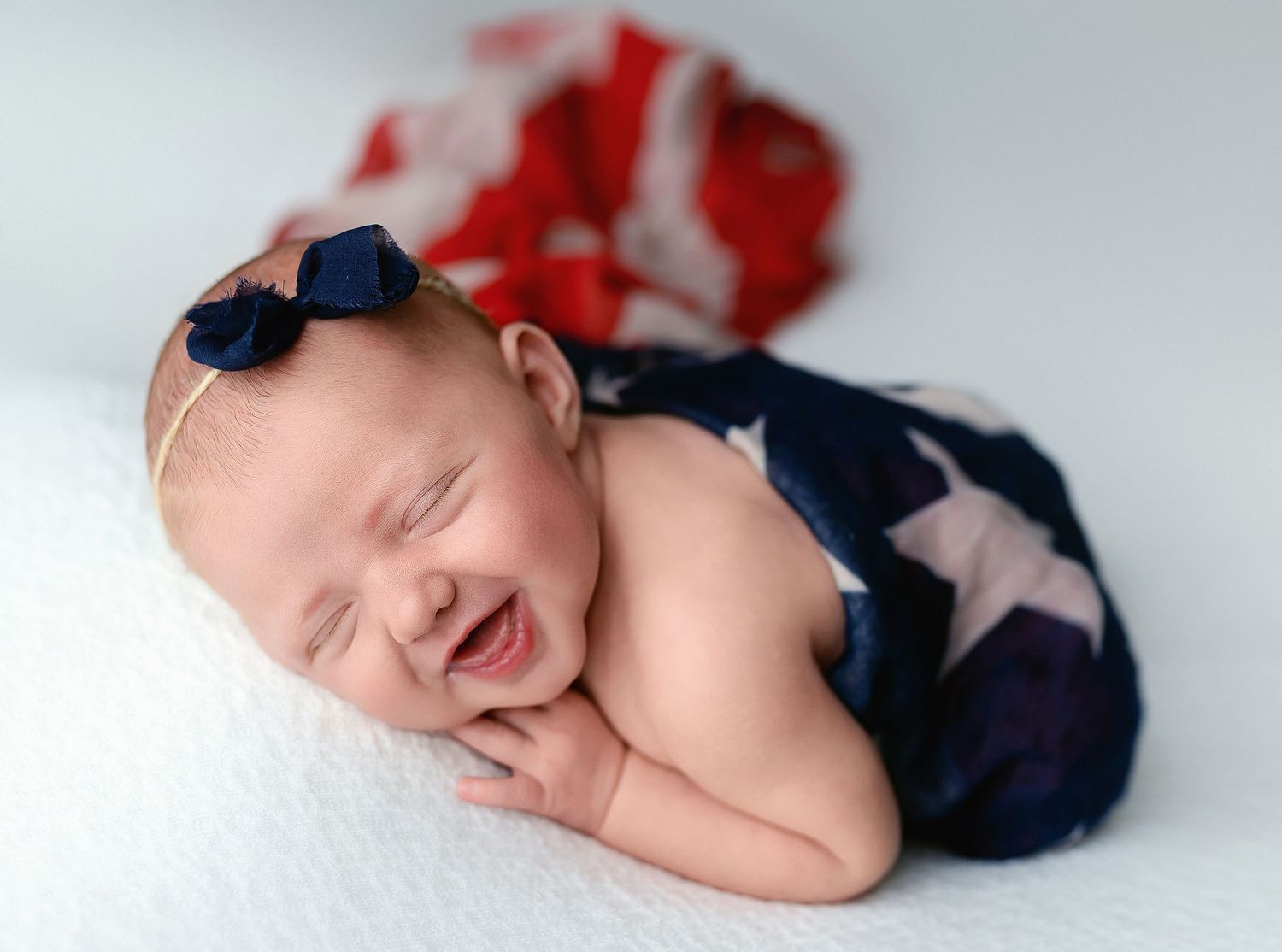 A newborn girl is laughing in the image, and she is wrapped in an American flag. - Greenville SC newborn photographer - Easley SC newborn
