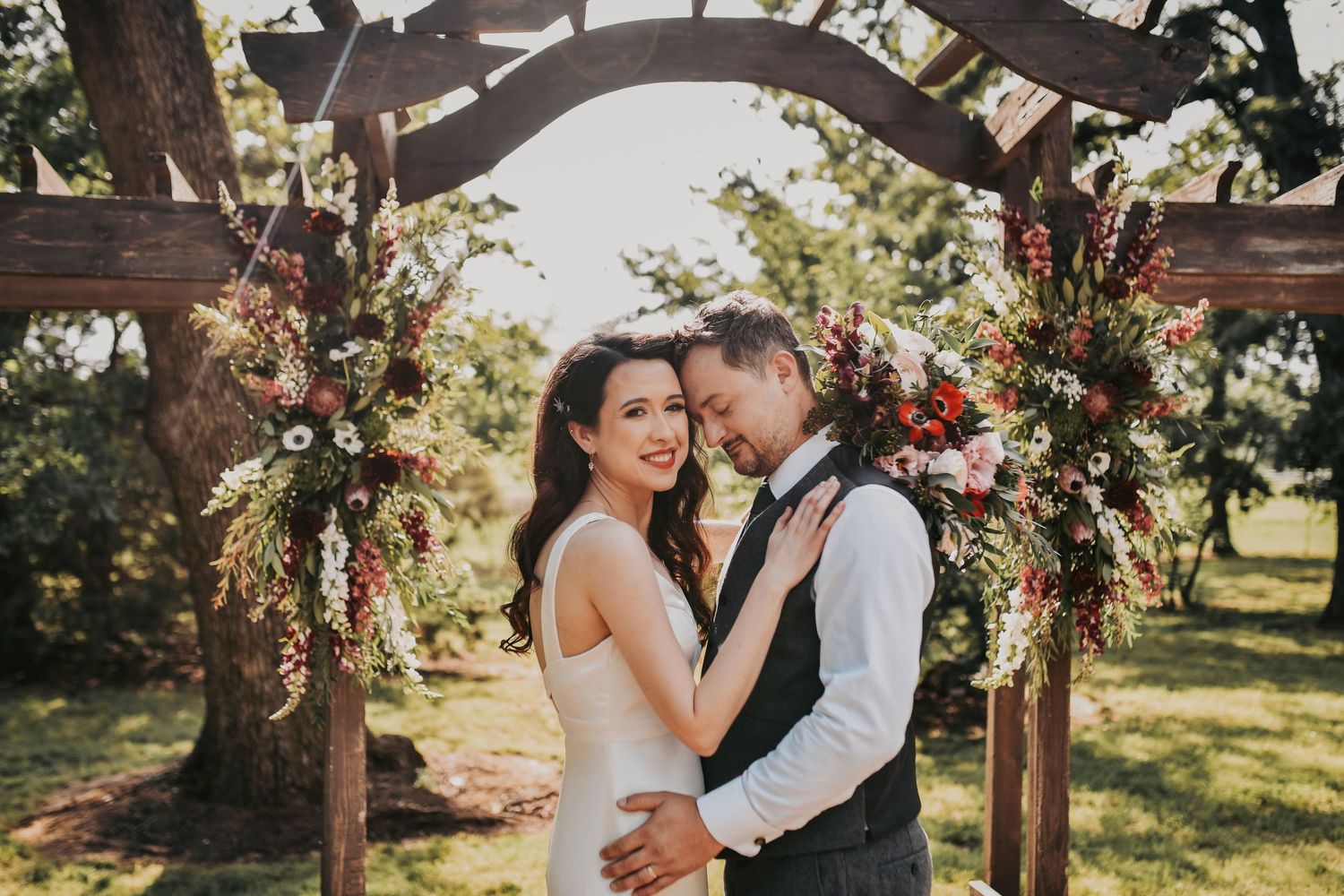 bride and groom embrace beneath oak trees at sunset in front of the wooden floral covered arbor at oak and ivy wedding venue