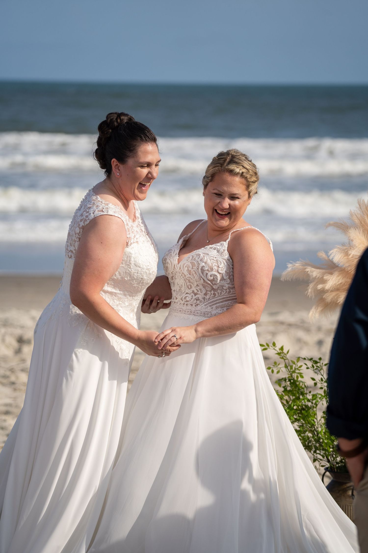 Two brides in white wedding dresses share a joyful moment together on a sunny beach.