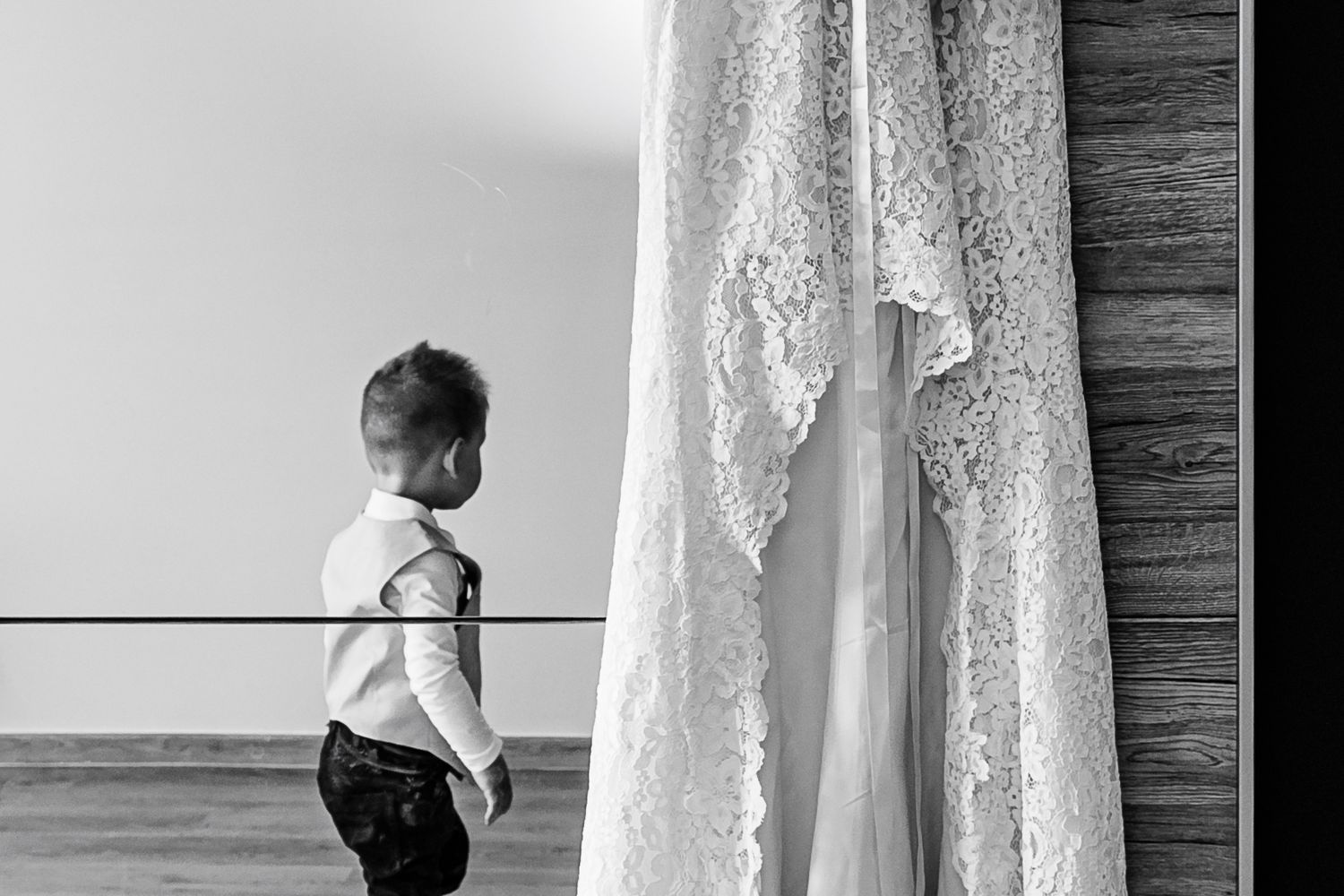 Black and white artistic image showing a delicate lace wedding dress hanging beside a wall while a child peers curiously.