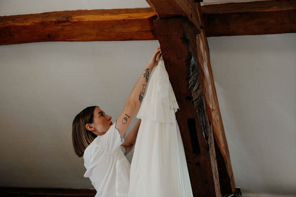 Woman with tattoos hanging a white wedding dress on rustic wooden beams in a barn venue.