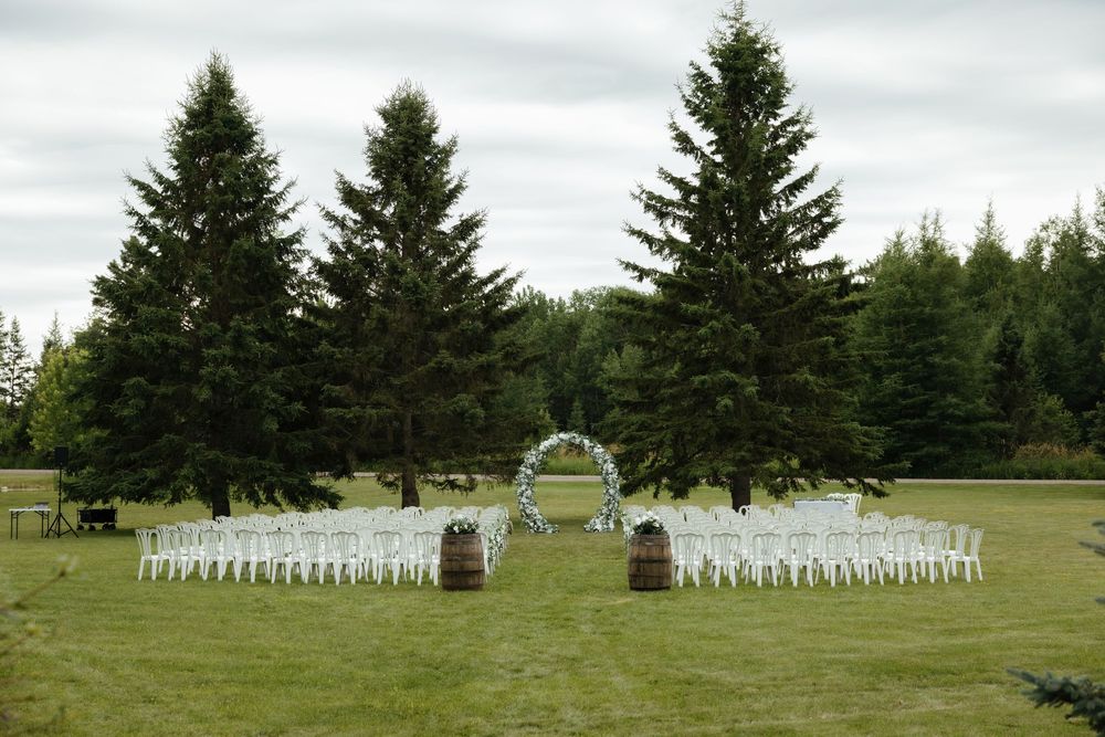 White chairs arranged in rows for an outdoor wedding ceremony with evergreen trees and an arch in the background.