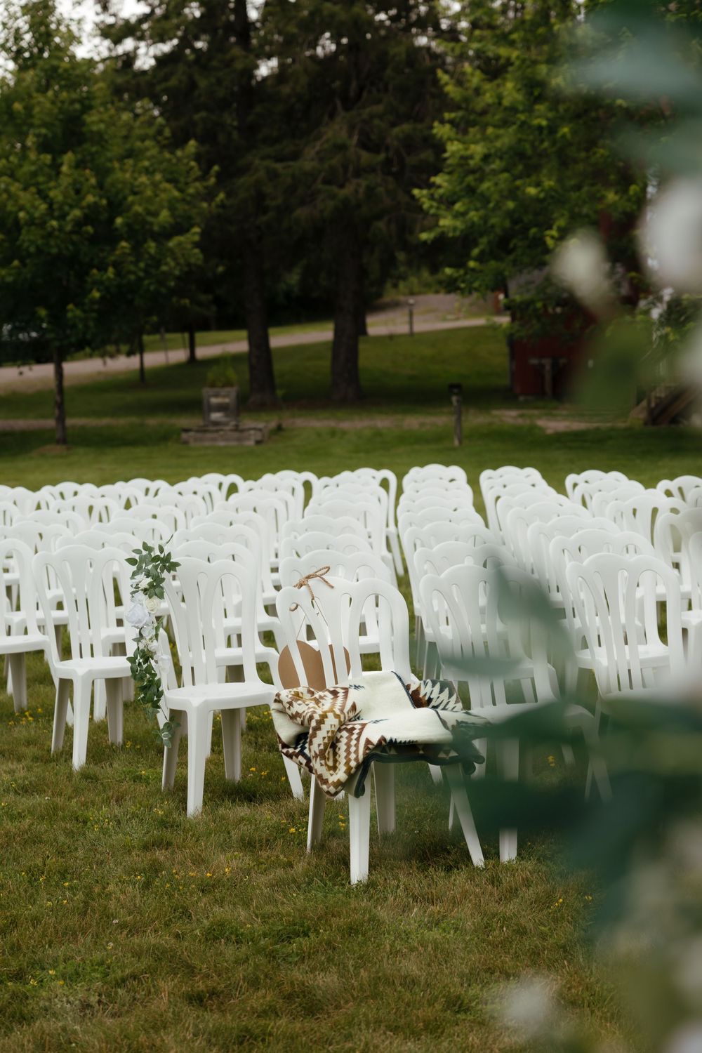 Rows of white plastic chairs set up on grass for an outdoor wedding ceremony beneath tall trees.