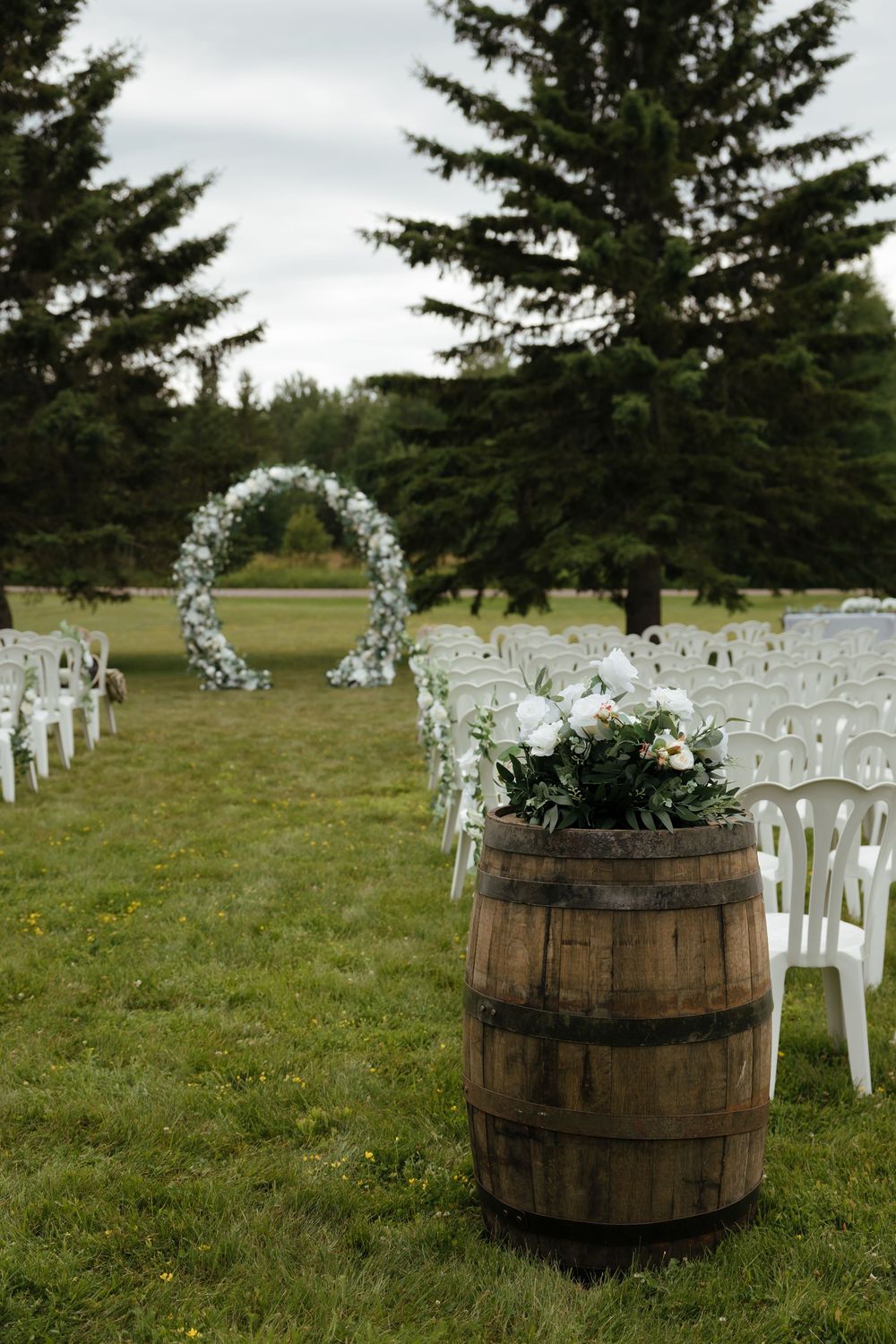 Rustic wooden barrels and white chairs arranged for outdoor wedding ceremony with evergreen trees in background.