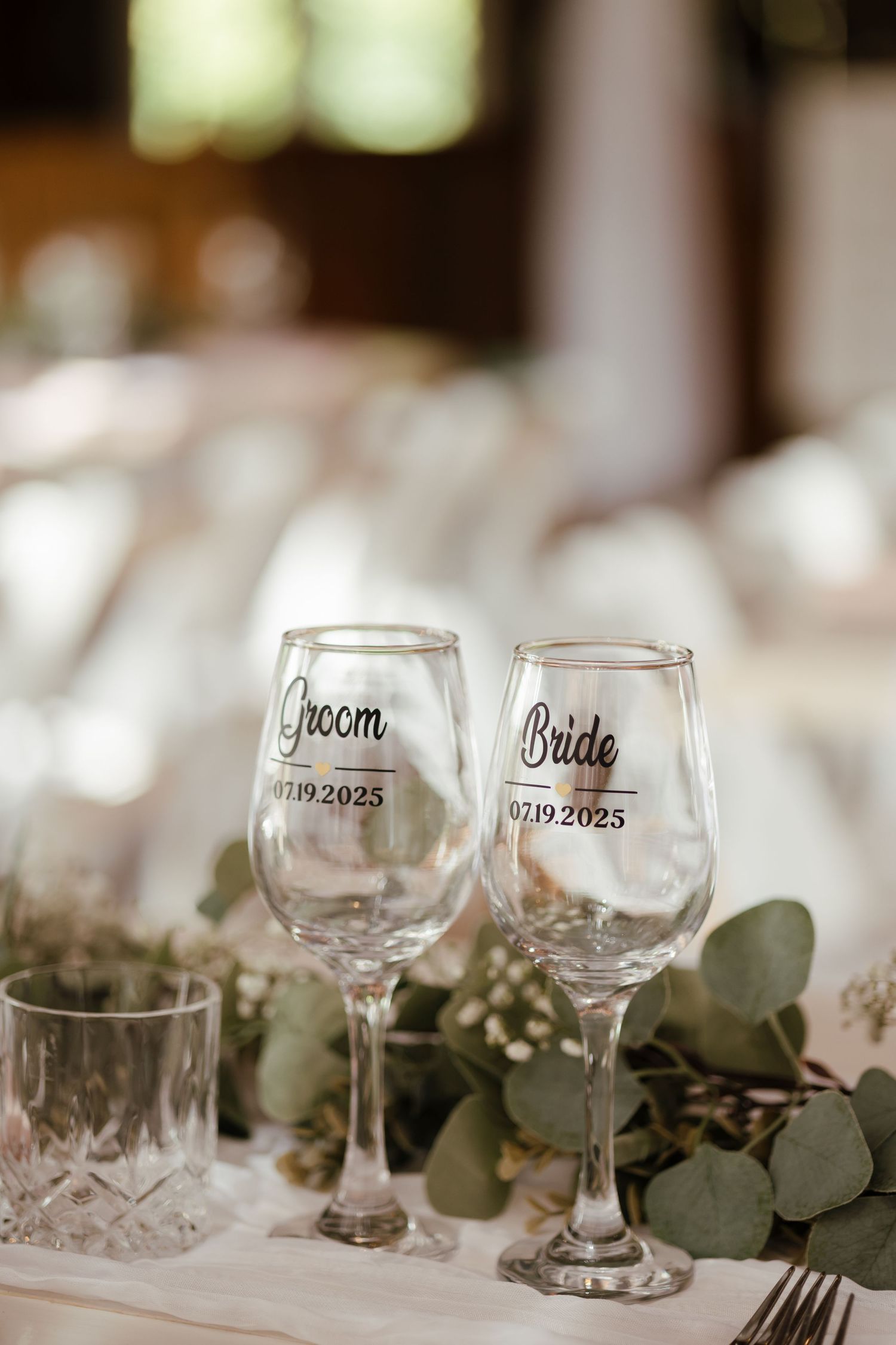 Wine glasses with custom text and eucalyptus leaves decorate an elegant wedding table setting.