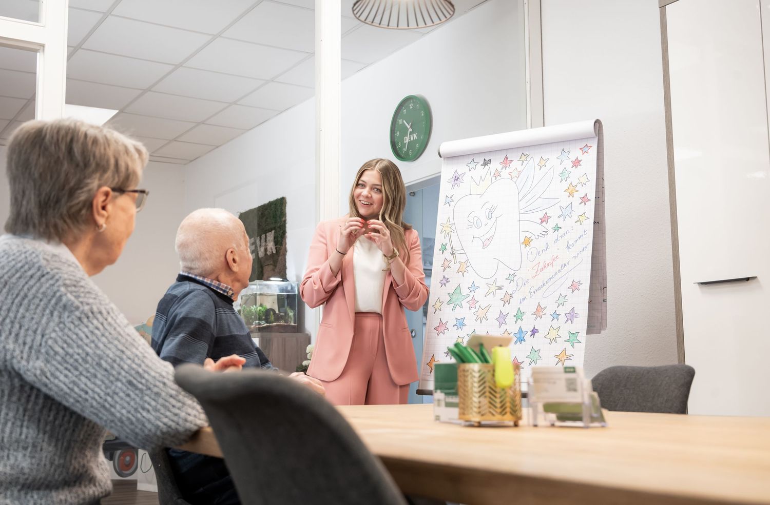 Eine Frau steht an einem white Board und erklärt einem Ehepaar etwas. Sie sieht man von vorne und das Ehepaar schaut sie an.
