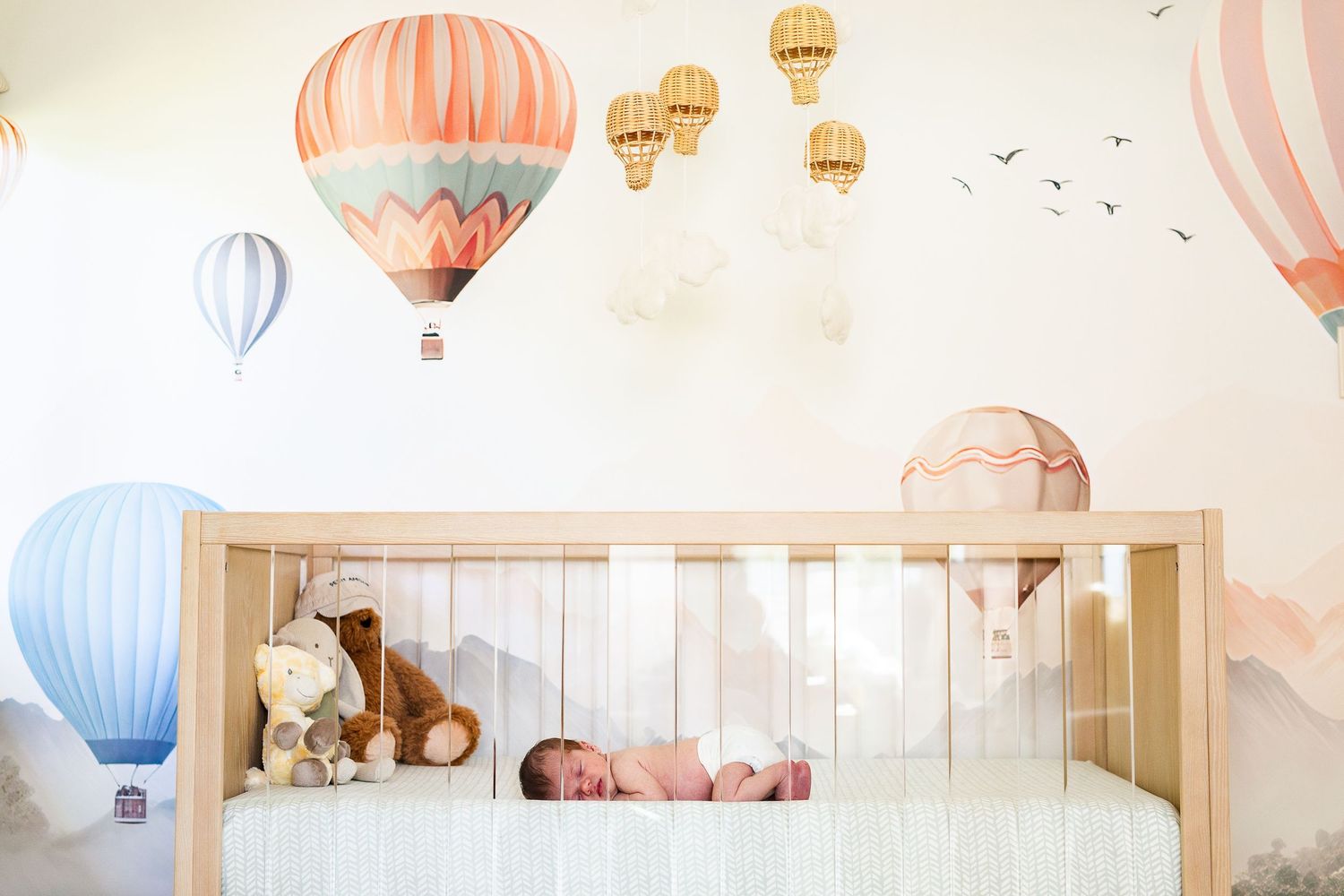 Charming nursery room decorated with hot air balloon wallpaper in pastel pink and blue tones with baby in crib.