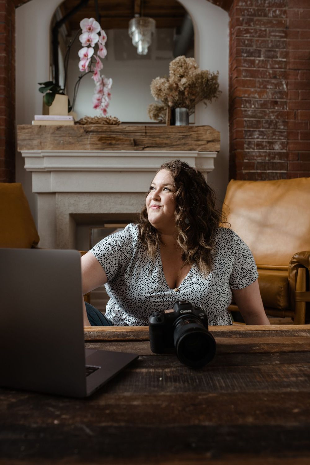 A cozy workspace setup with laptop and camera on wooden table near fireplace with dried flower arrangements.