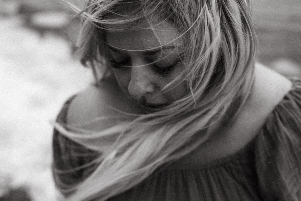 A black and white close-up photo shows windblown blonde hair framing a downward-turned profile in soft focus.