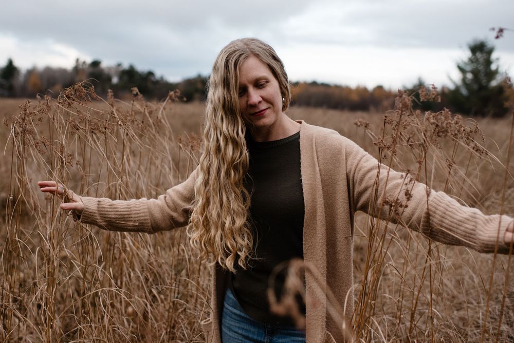 A carefree person with long hair wearing a beige cardigan stands with outstretched arms in a field of tall autumn grass.