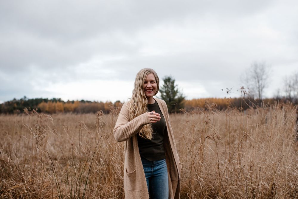 Woman in light brown sweater walks through a golden fall field of tall grass at sunset.