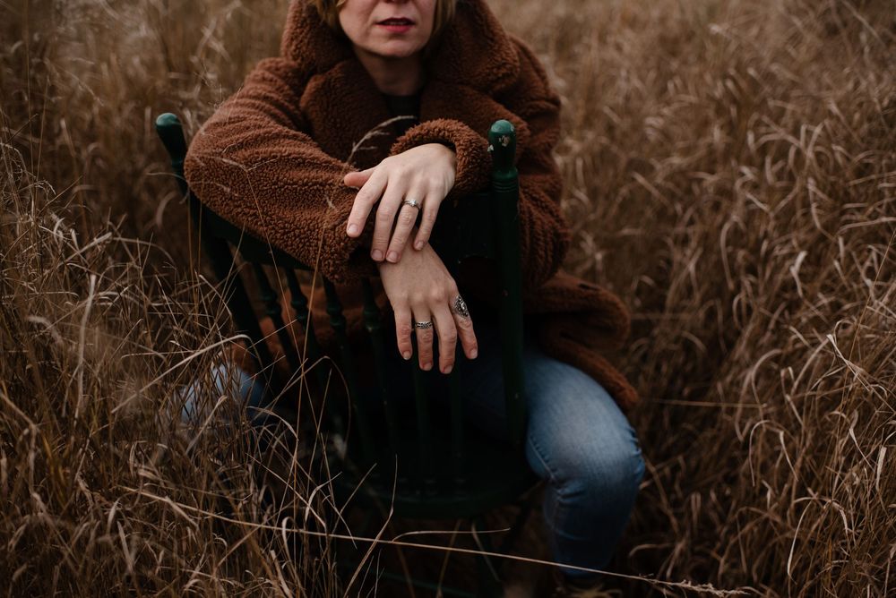 A moody autumn portrait showing someone wearing a brown knit sweater seated among tall dried grasses.