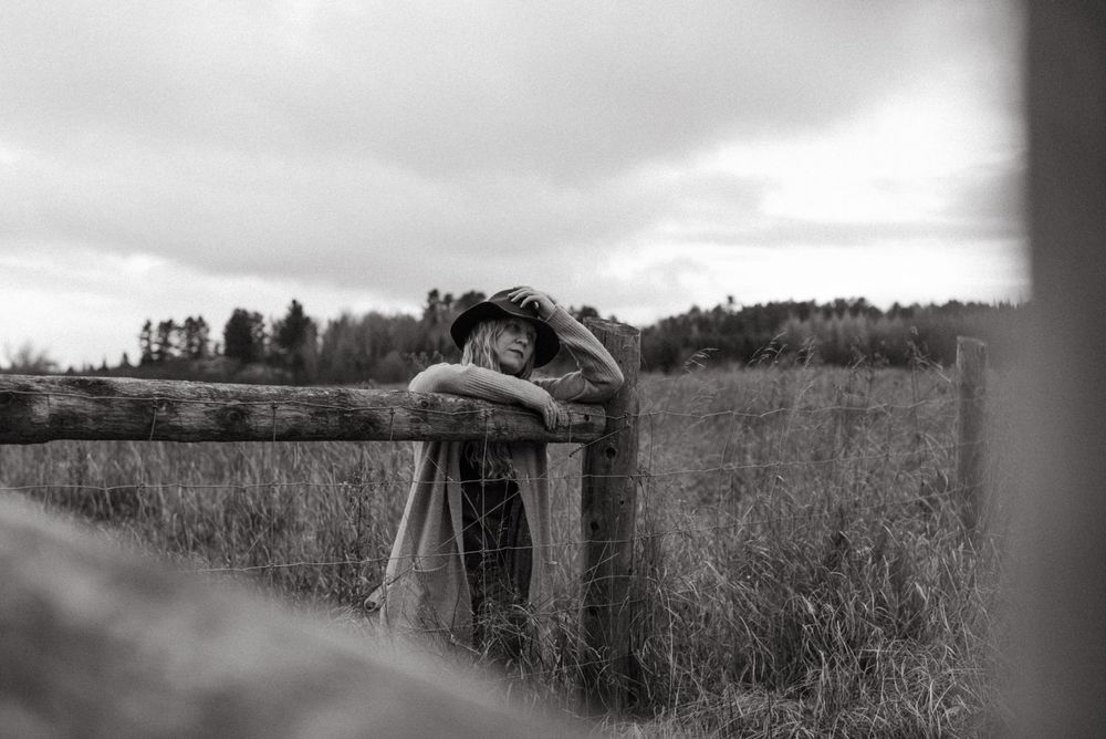 A black and white artistic photograph shows someone resting on a wooden fence post in a rural meadow setting.